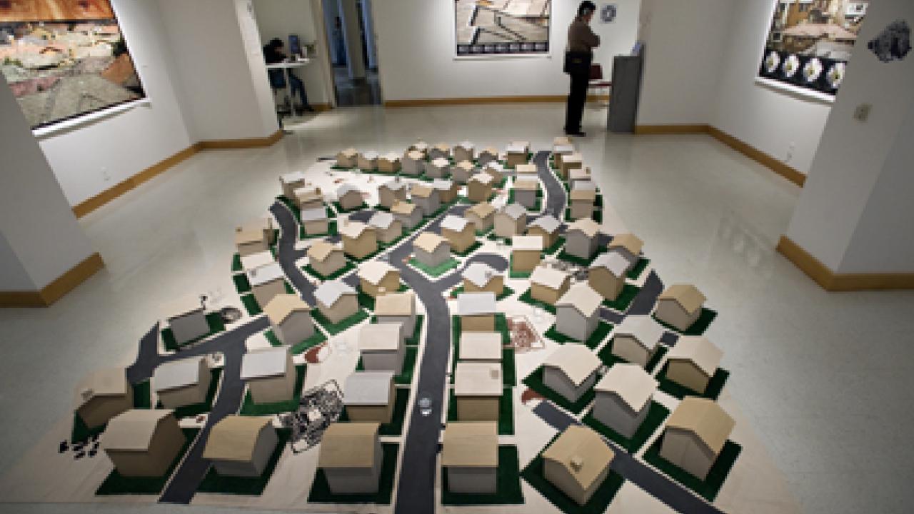 Floor installation of miniature houses on patches of green lawn along suburban streets, all on top of a map depicting American Indian symbols.