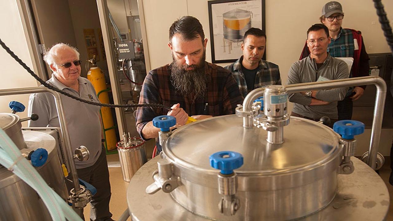 Four men watch another man working with a stainless steel vat