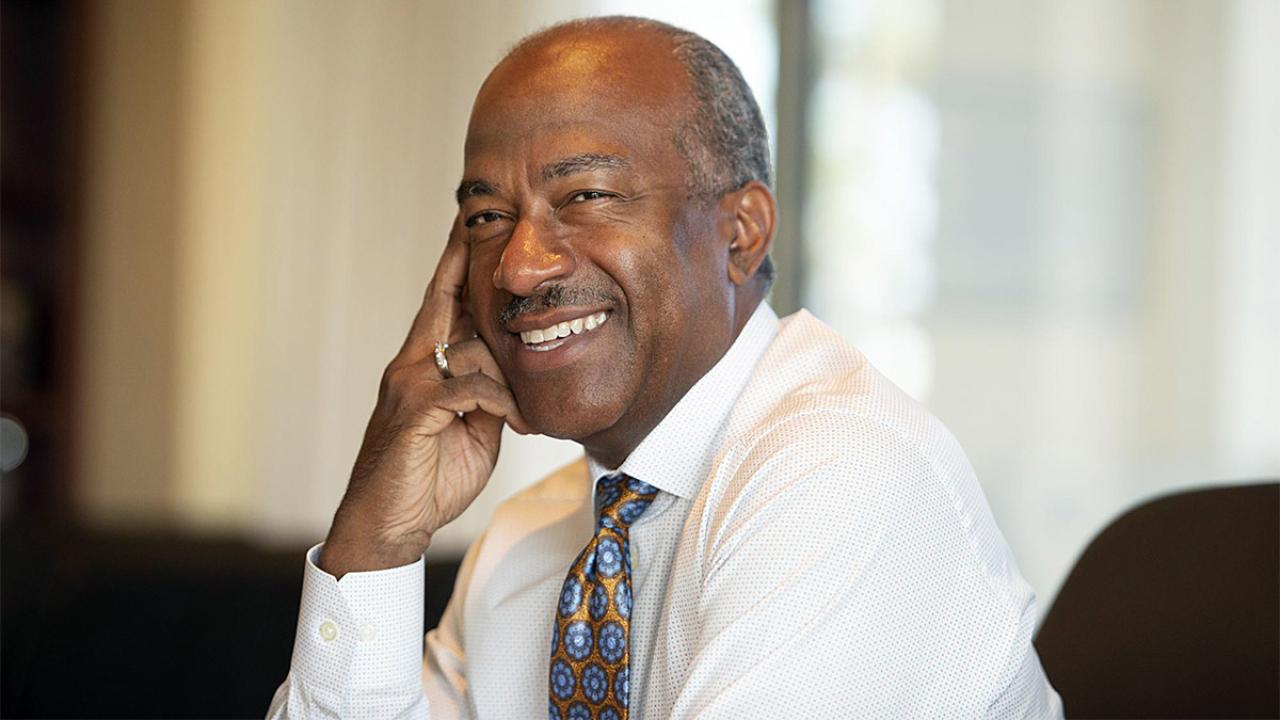 Chancellor Gary S. May smiles, sitting at desk.