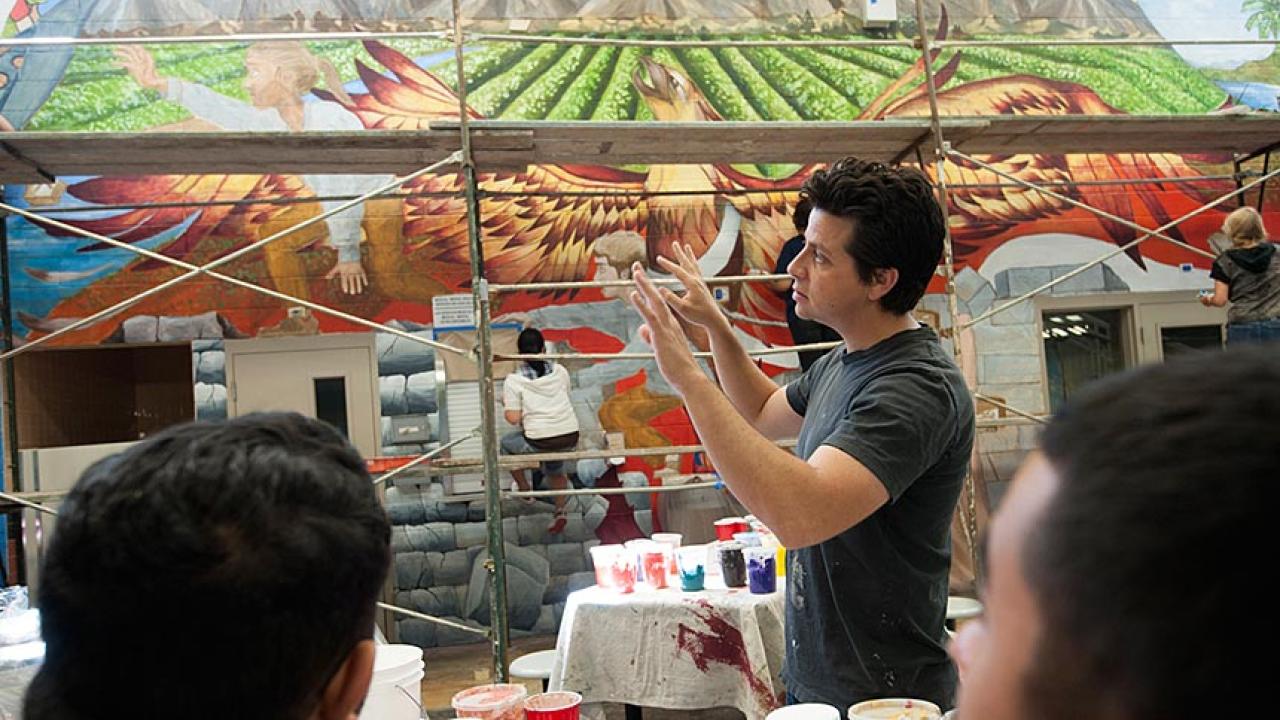 Man gesturing in front of a painted mural with two people watching him