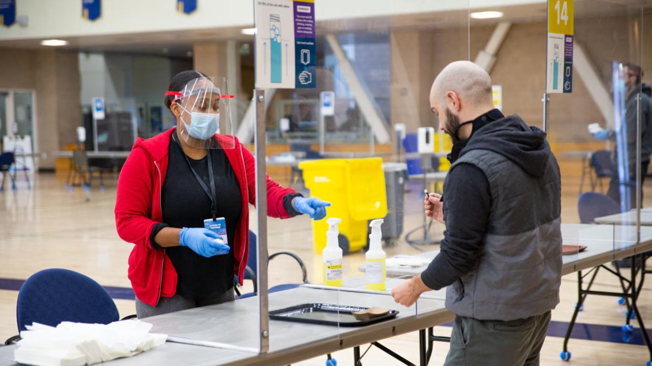 Woman behind plastic shield directs man at testing kiosk.
