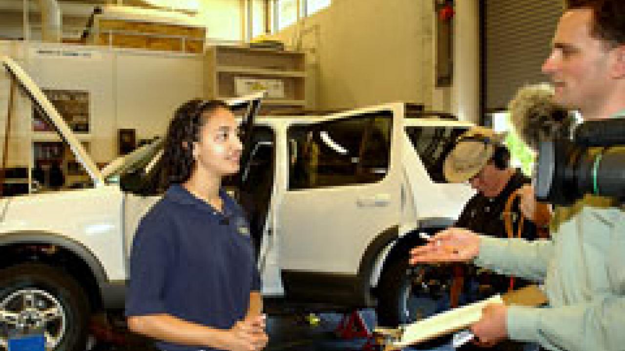 Man on right interviewing woman with Ford Explorer in the background with the hood up.