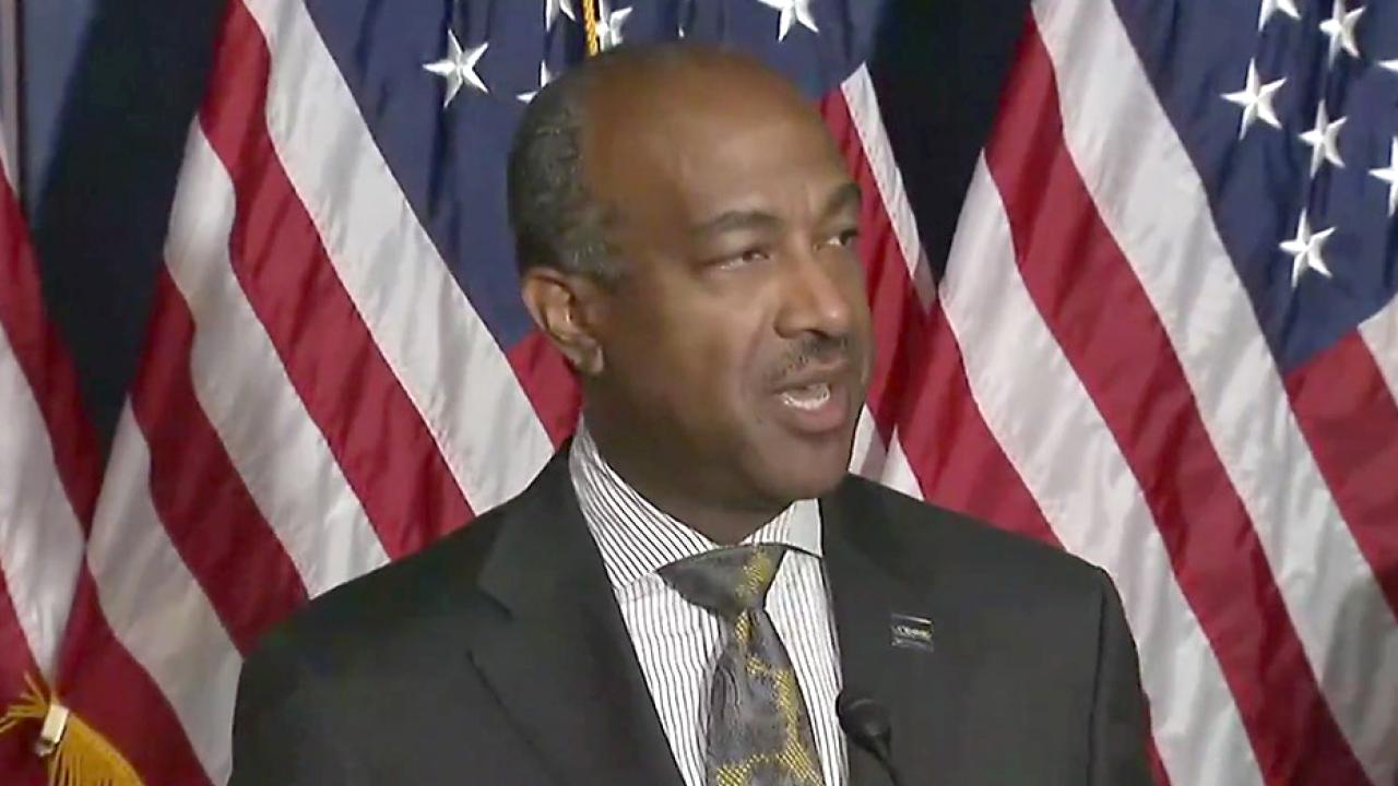 Chancellor May at podium, with backdrop of U.S. flags