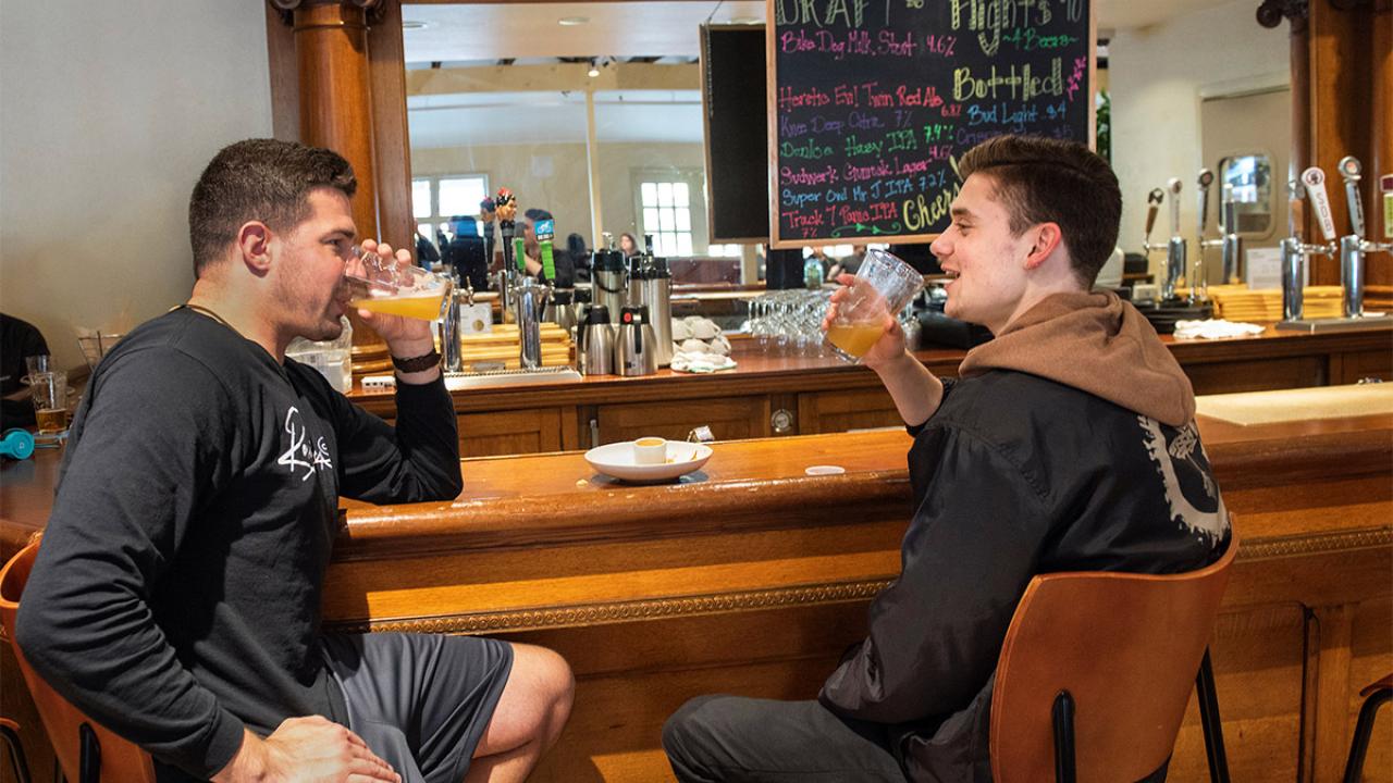 Two students drink beer at a wooden bar.