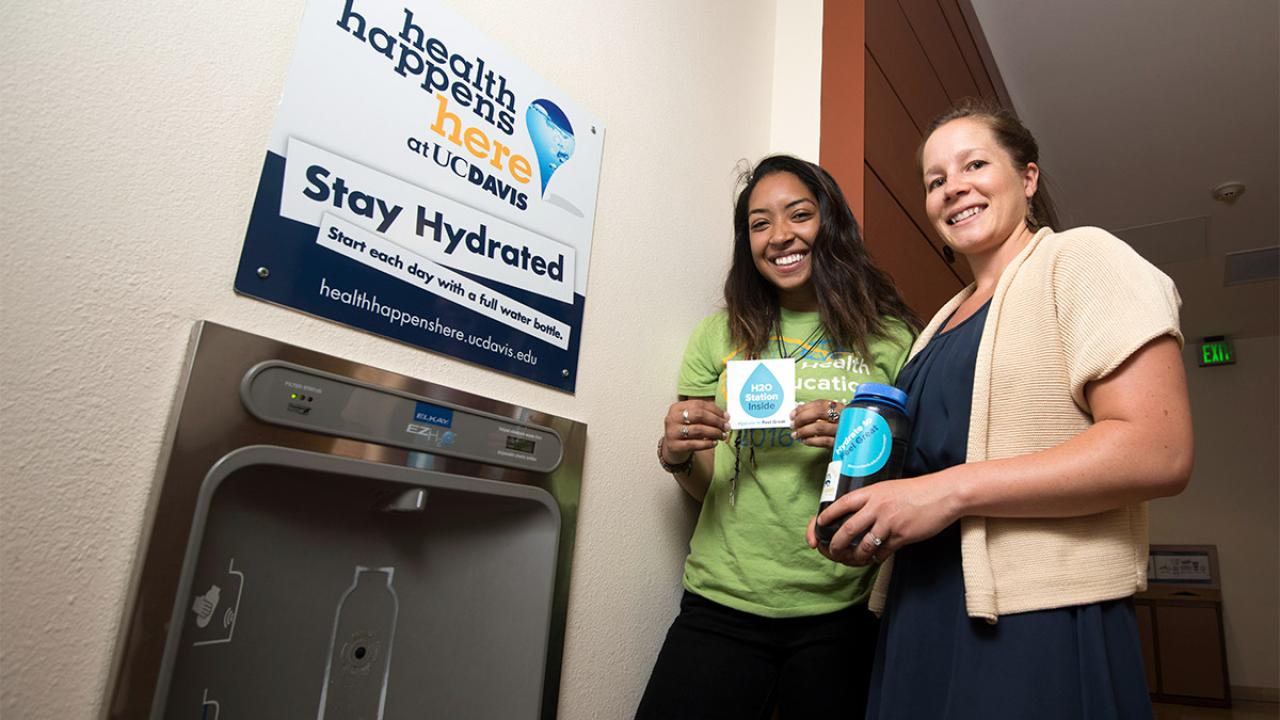 Two women stand next to station for filling water bottles.