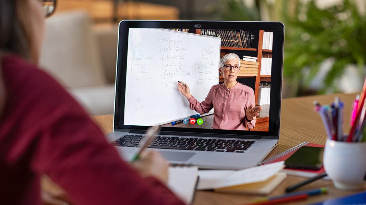Instructor at whiteboard, on monitor, with a woman sitting at the monitor, taking notes.