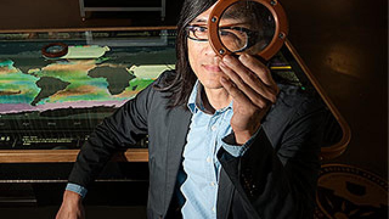Man sitting at a glass table holding an magnifying circle over his eye