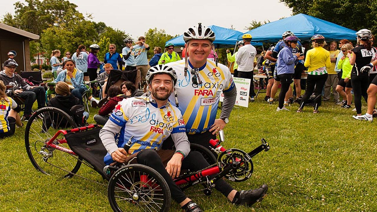 Man in bike helmet and jersey kneels next to a man seated on a recumbent bike with people and event tents behind them