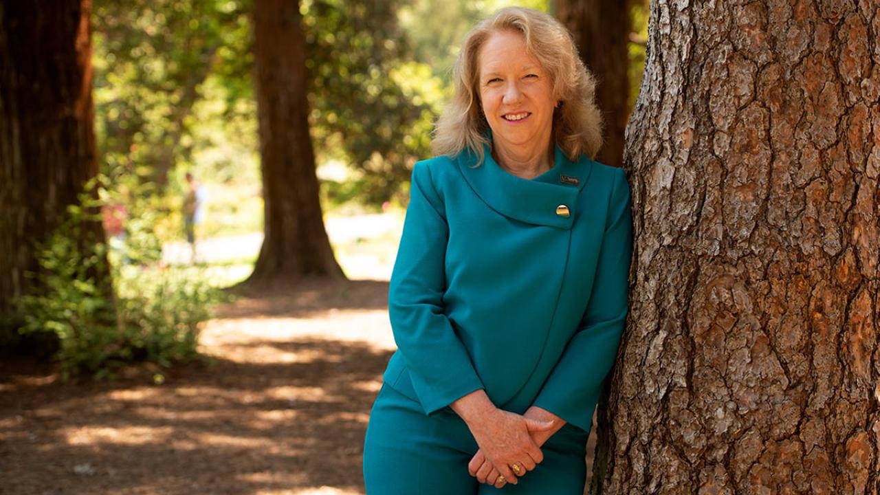 Provost and EVC Mary Croughan leans against tree in Arboretum.