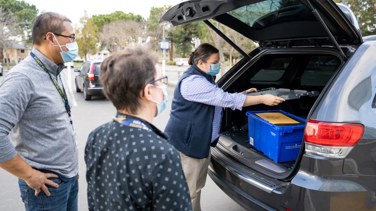 Two people watch as another psreon unloads blue box from vehicle.
