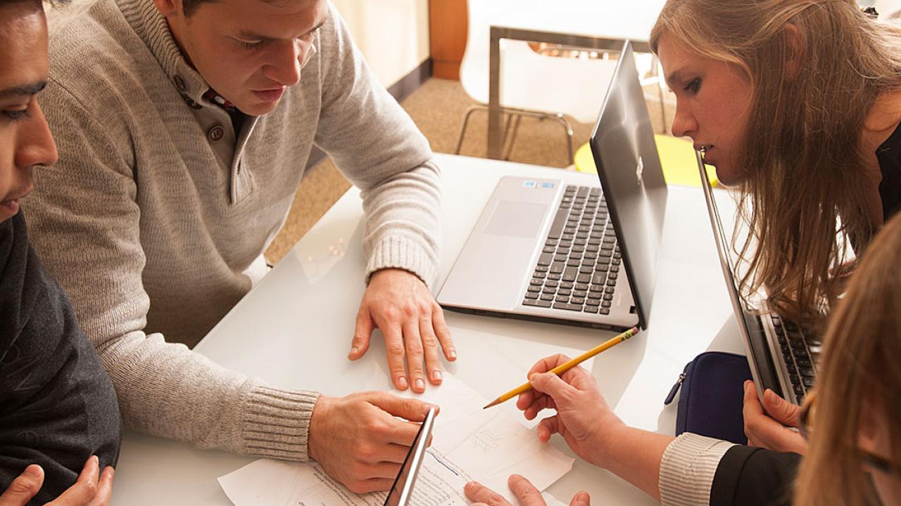 Three students intent on a drawing on a table as a person shown by only the top of her head draws