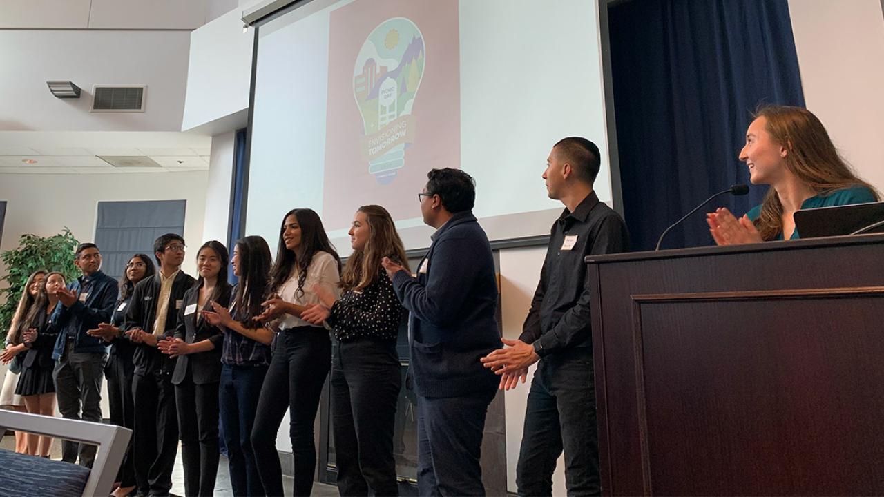 Students stand in a line and clap while looking at a projector screen.