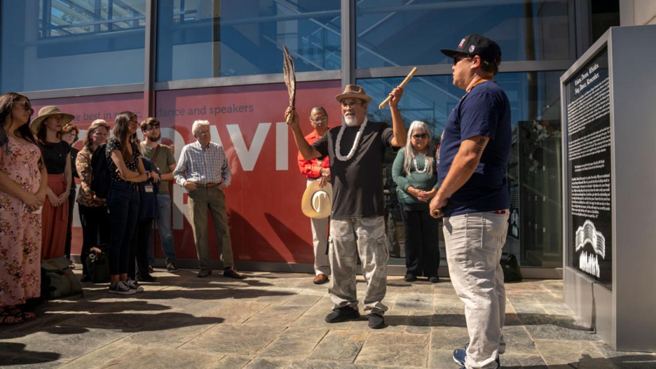 Man performs Indioan blessing while holding a clapper (stick) and feathers, in front of new historical marker.