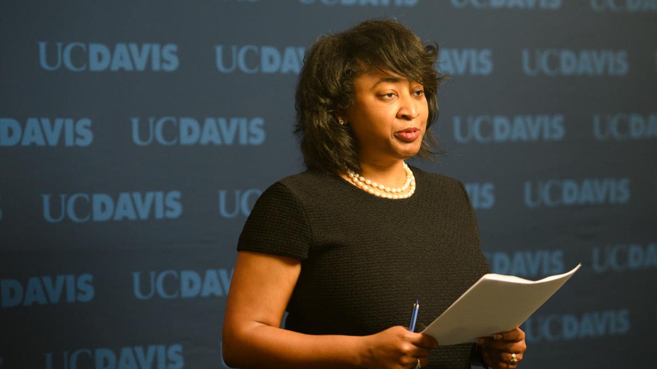 Woman reads from script against "UC Davis" backdrop.