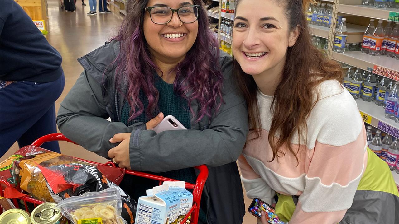 Two women stand next to full grocery cart.