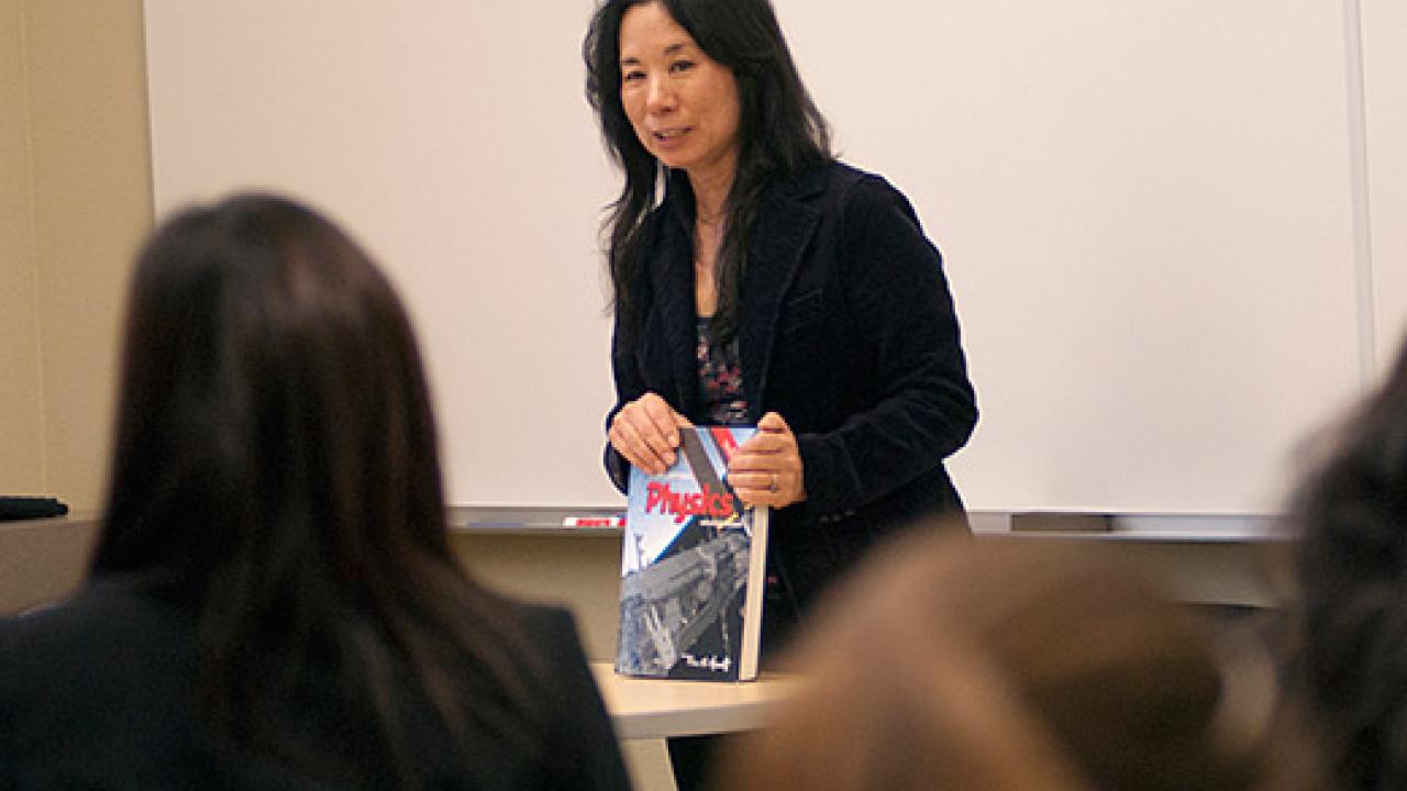 Photo: female teacher addressing her class and holding a physics book