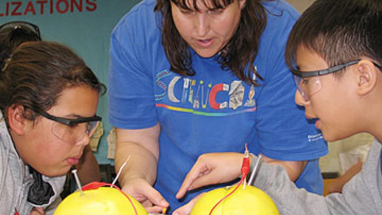 Photo: two young students and a woman examine two grapefruits set up to conduct energy