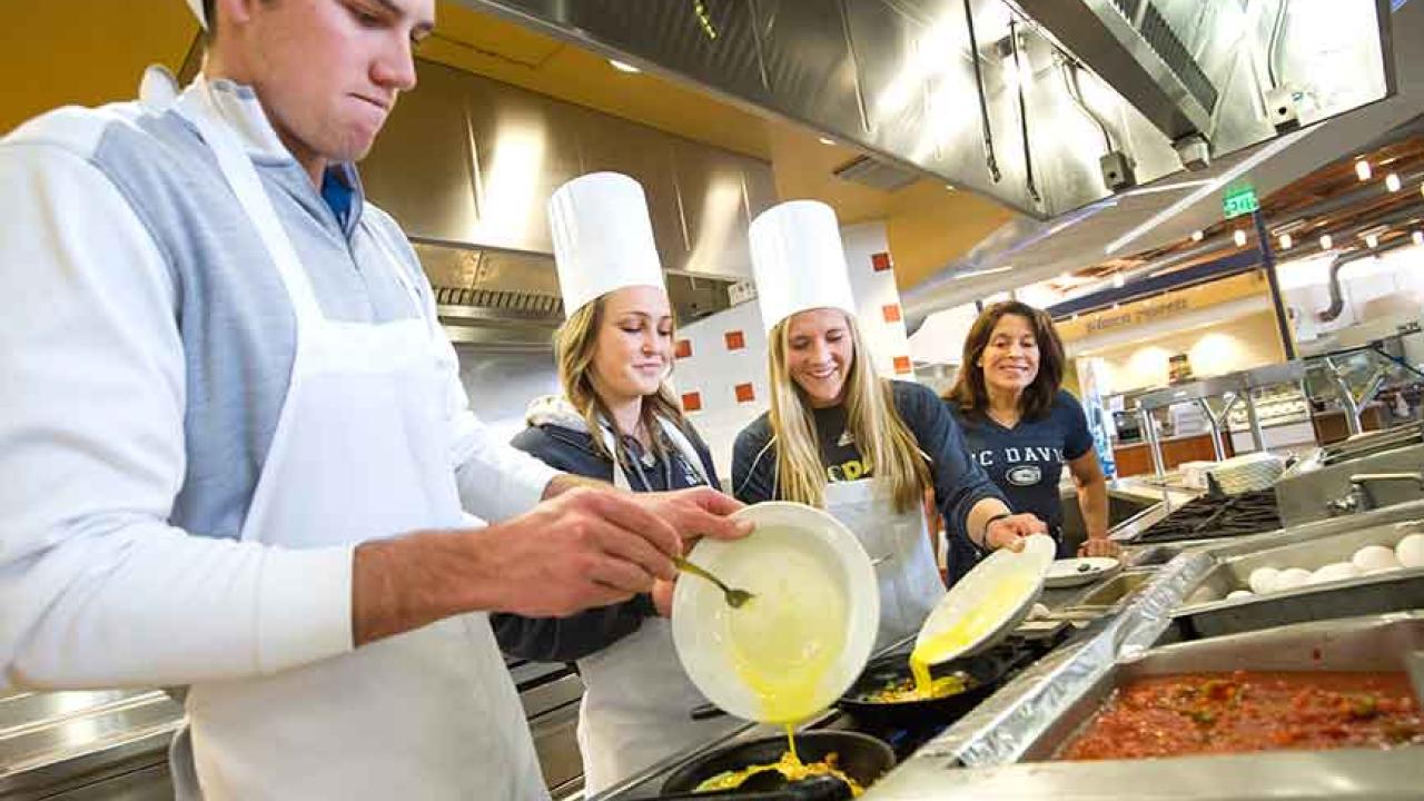 Photo: Students pour eggs into scrambler dishes, at a stove in the dining commons.