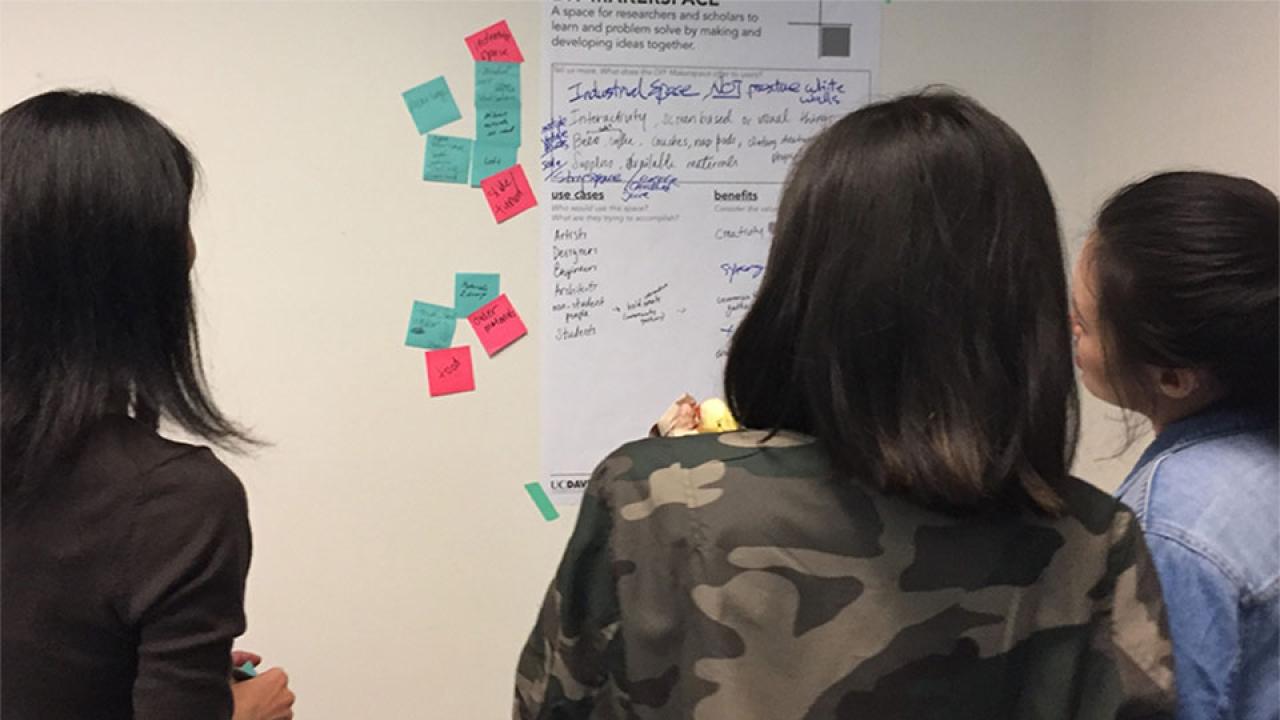 Three women look at wall display with small notes around it.