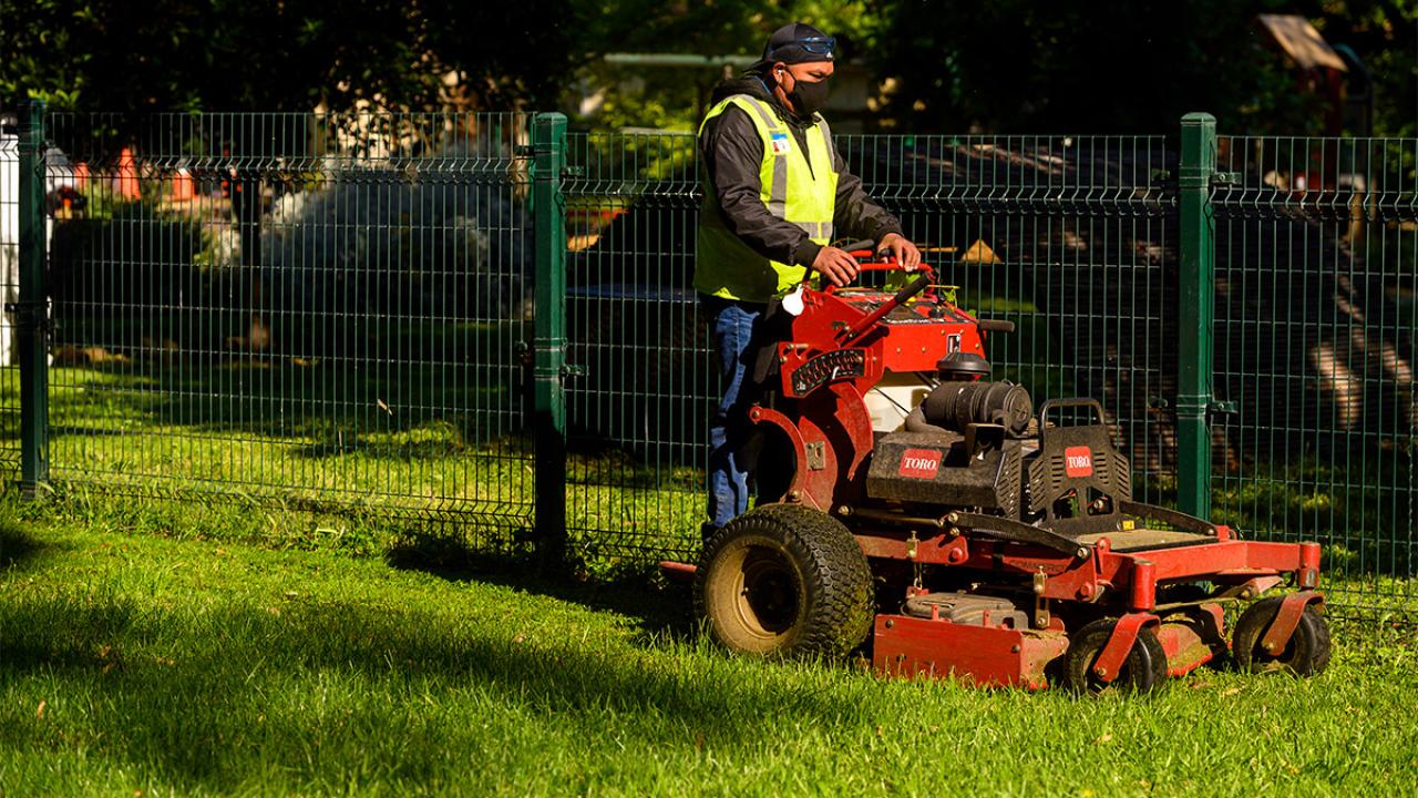 Man wearing mask stands on lawn mower.