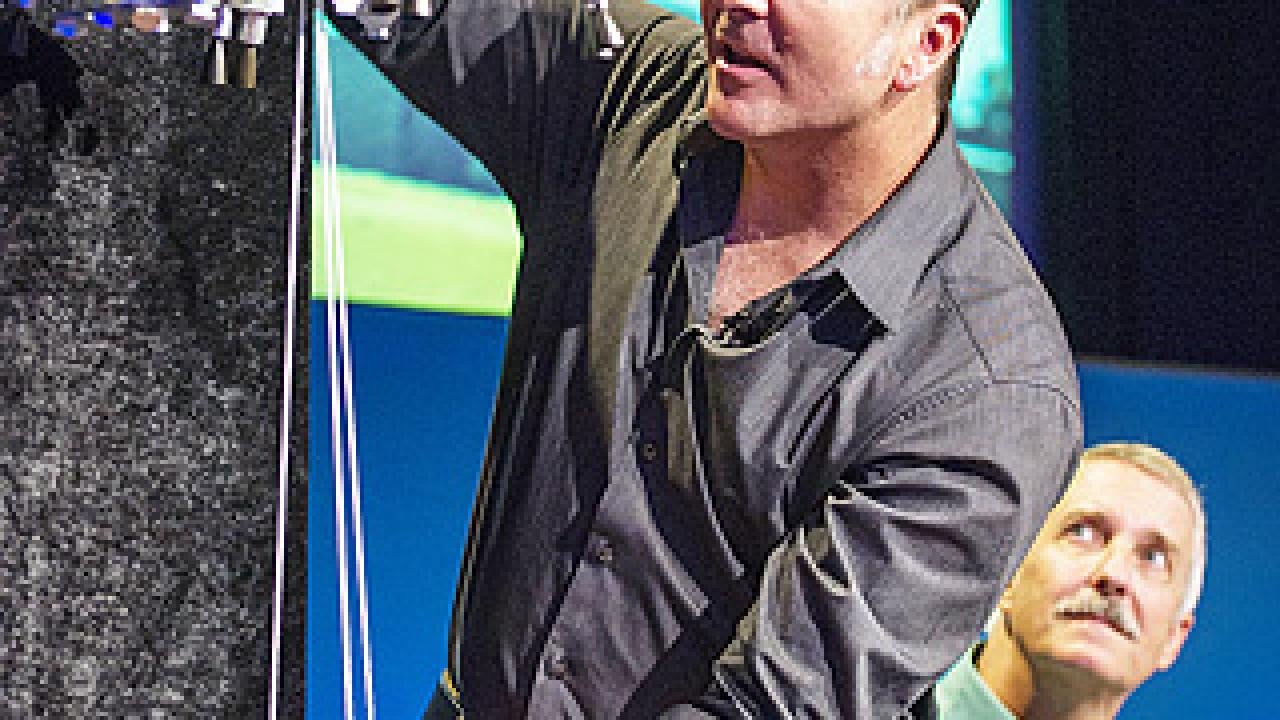 Man demonstrating Mars rover on strings lowering to ground, as another man in background looks on