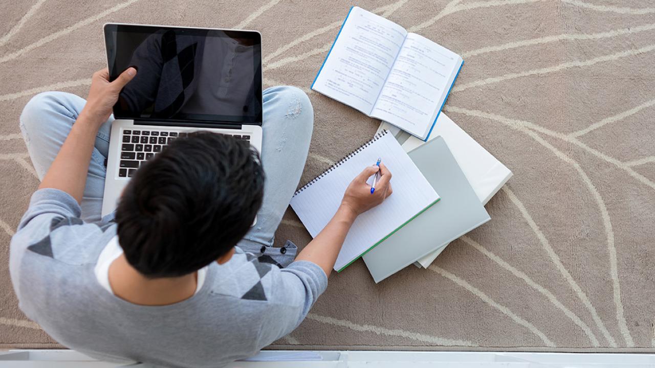 A student sits on the floor with a laptop and notebooks.
