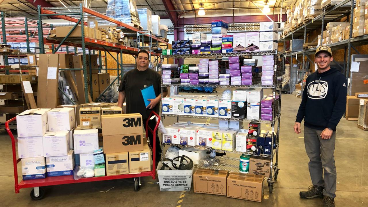 Two men stand amid stacks of supplies.