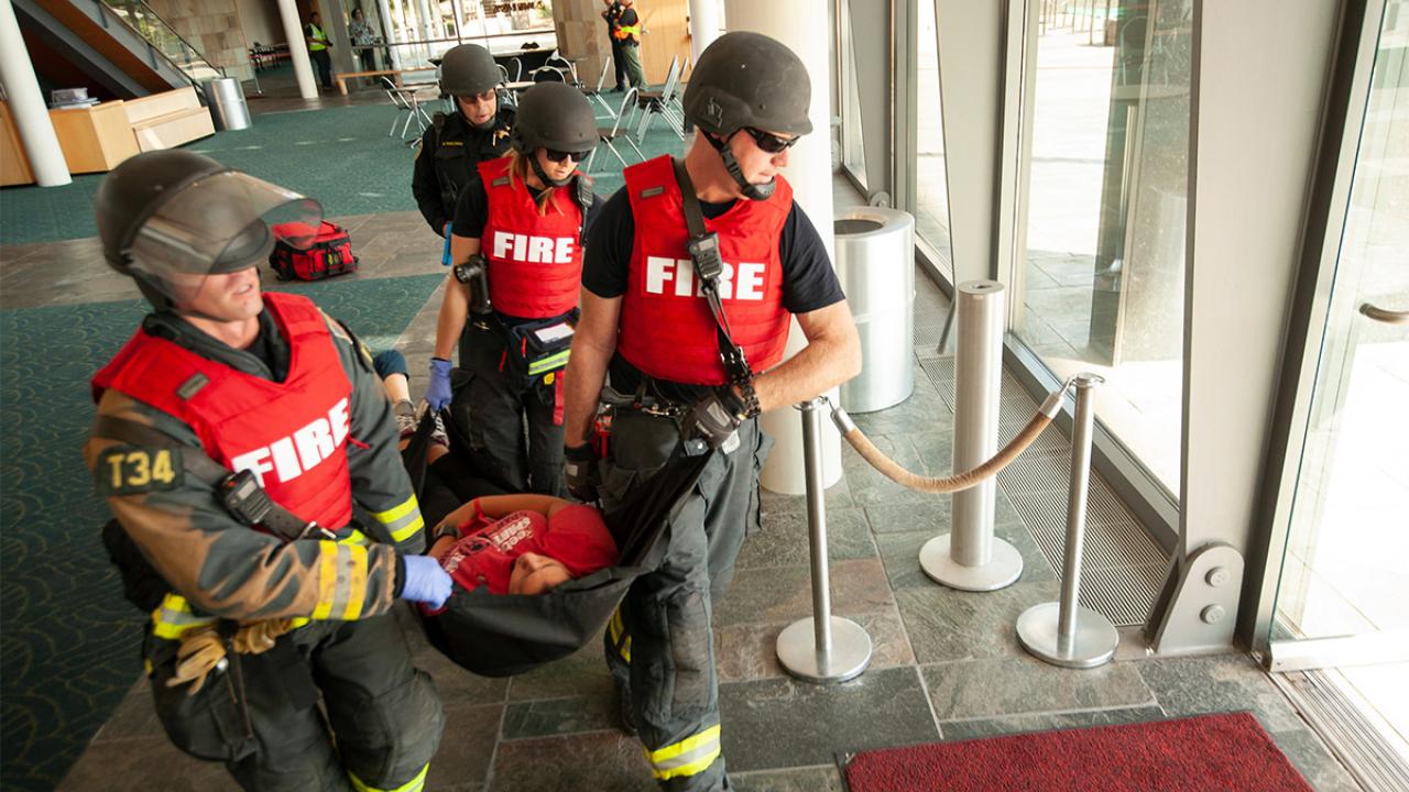 Three firefighters in helmets and red vests carry a patient in a stretcher, followed by a police officer