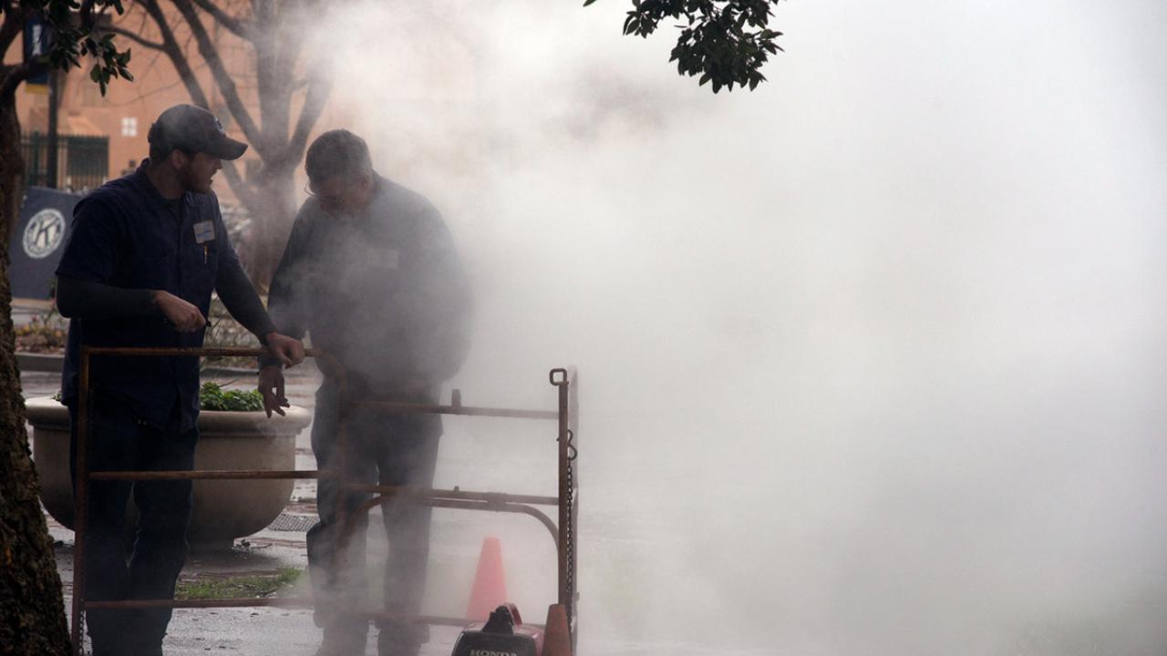 Two workers stand over a grate emitting steam.