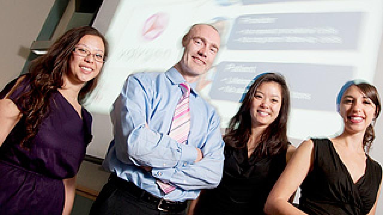 Four people, a woman, a man and two women posing in front of a PowerPoint screen in the background