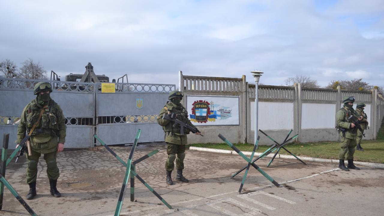 Four armed men in green uniforms, helmets and goggles stand in front of a concrete fence. 