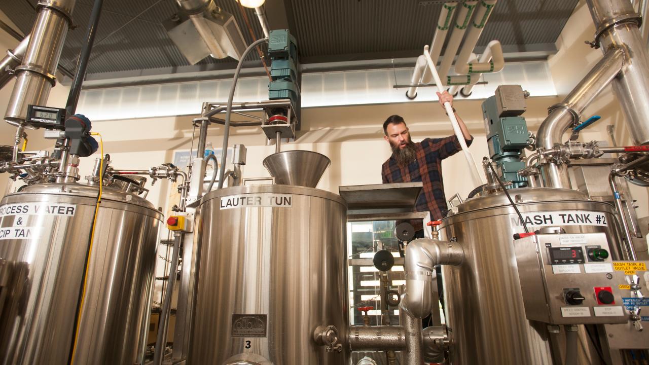 A man with a long beard and plaid shirt stands behind a row of silver metal tanks joined by pipes. 