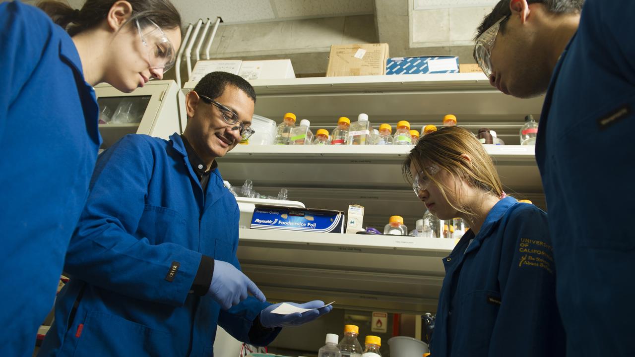 Four people in dark blue labcoats and protective glasses stand in a half-circle in front of a laboratory bench. 