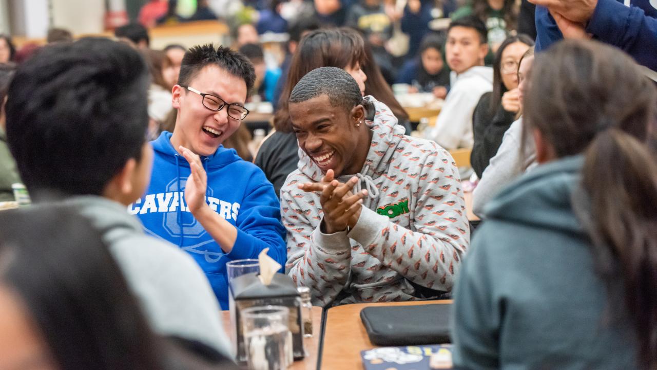 amidst a full indoor dining common, two students in the center frame of the photo are laughing next to each other. One is wearing glasses with a blue hoodie while another wears a grey top. 