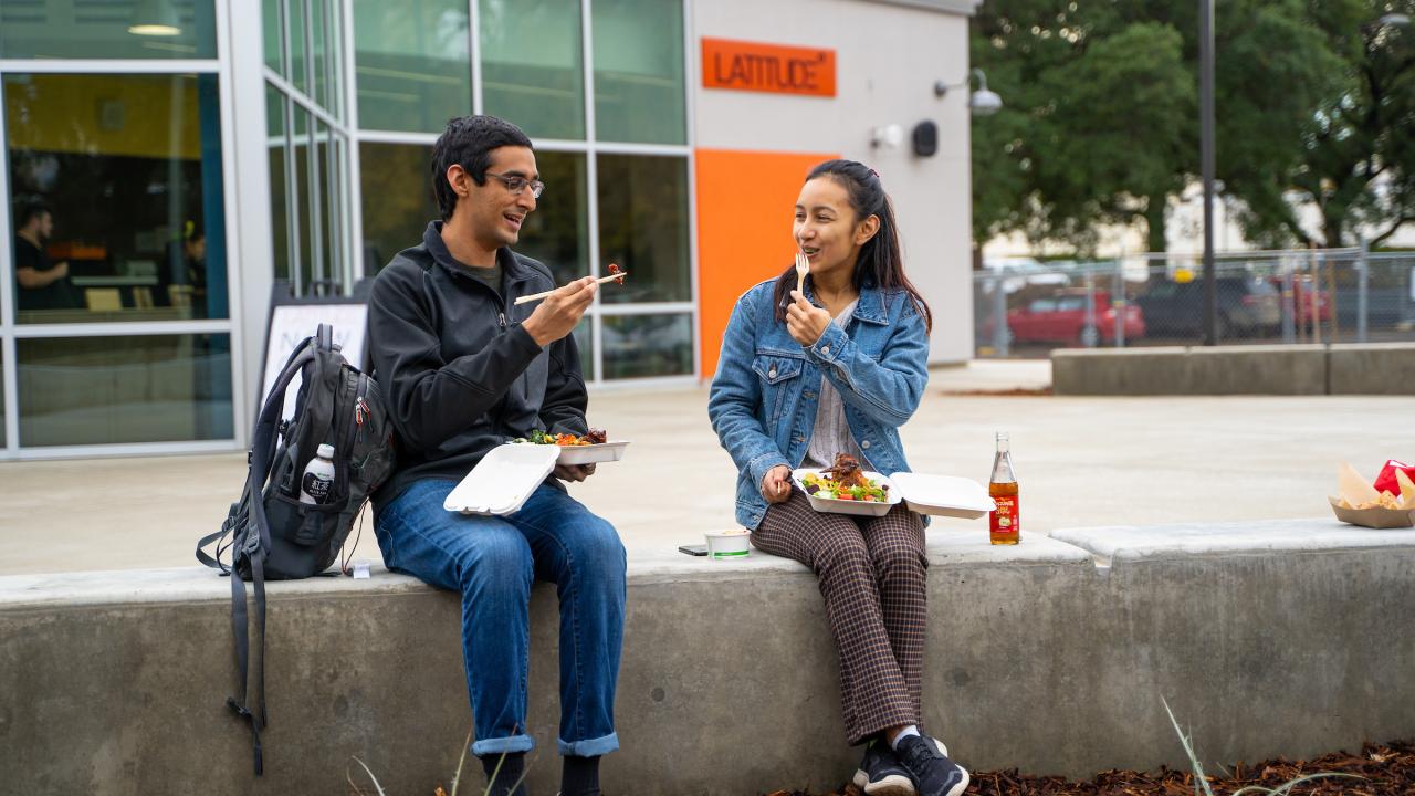Two students sit outside Latitude Dining Commons eating food with chopsticks. They are facing the camera and angled towards each other on a sunny day. 