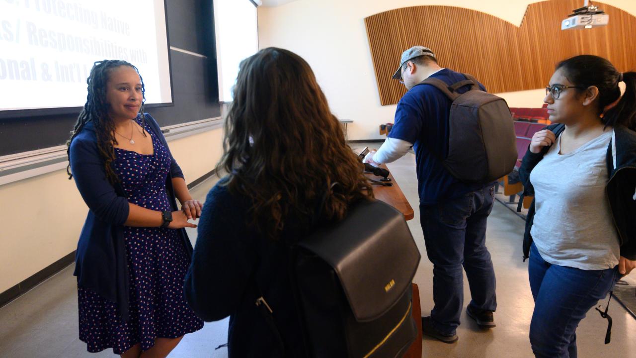 Woman professor talks to students after class with whiteboard in back.