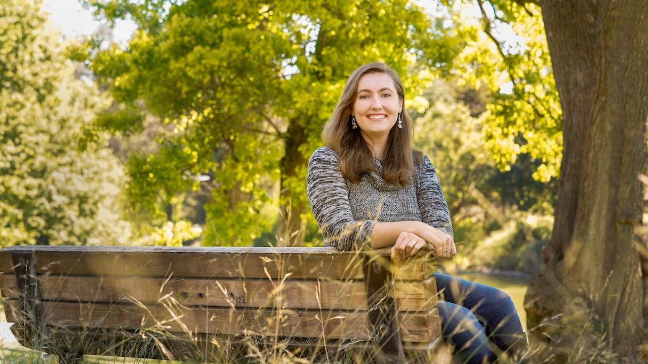 Woman sitting on park bench smiling at camera with lake and trees in background