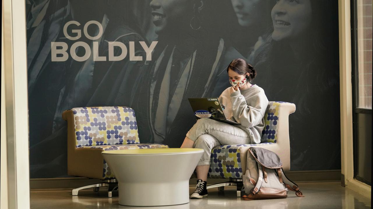 The words Go Boldly appear on a wall behind a young woman studying on a laptop. 