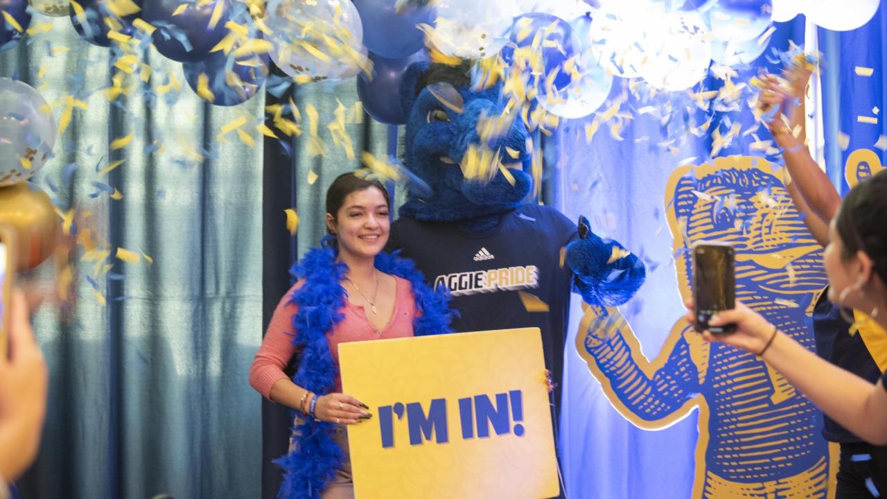 Student wears a blue feather boa poses with Gunrock during an Aggie Day celebration, holding a yellow “I’m In!” sign as blue and gold confetti falls.