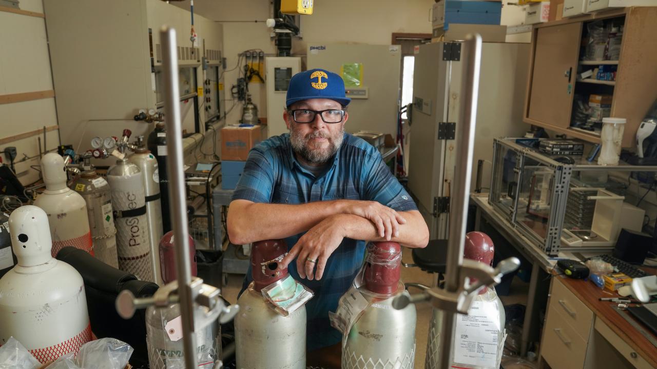Man behind scientific equipment in lab looking at camera in blue ball cap 