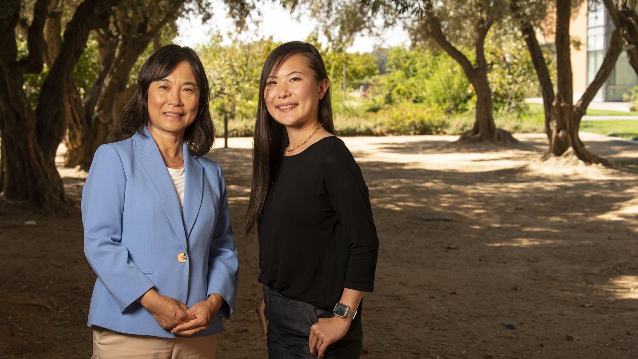 Two women photographed outdoors. The woman on left is wearing a blue shirt and facing the camera, a younger woman to the right is side on with her head turned towards the camera. 