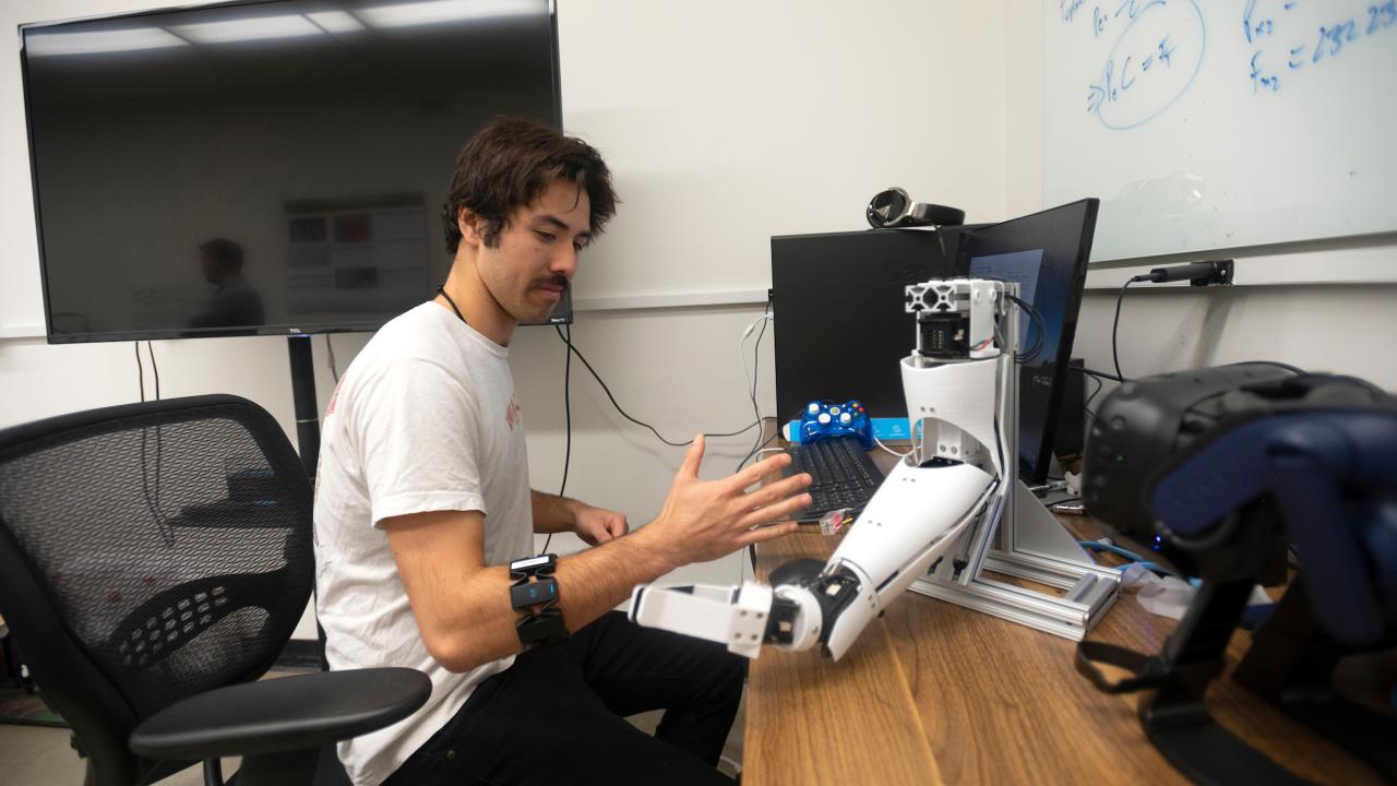 A young man in a white t-shirt sits at a desk and flexes his forearm, which has a band around it. On the desk is a robotic arm and a computer monitor. 
