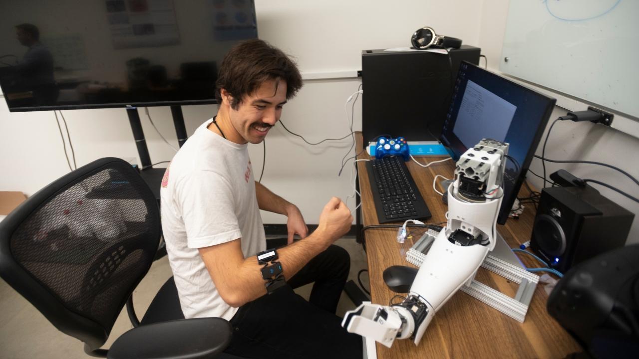 A person sits at a desk with a robotic arm, smiling and making a fist.