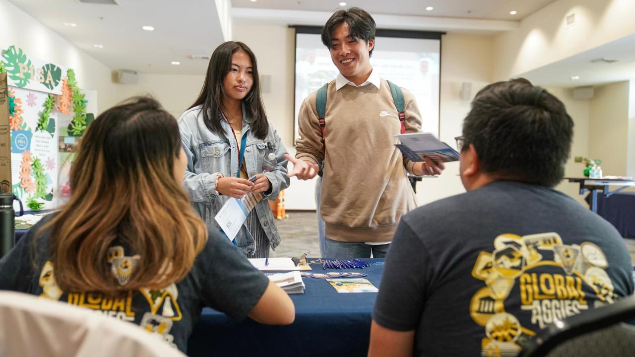 Two students, one in a tan sweater and the other in a jean jacket, stand in front of a table and ask questions to two UC Davis representatives in navy short sleeve Global Aggie shirts.  
