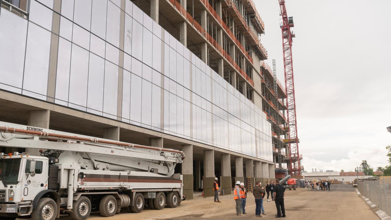 A large building under construction with open concrete beams visible above a glass wall. People wearing hard hats stand in the middle distance. 