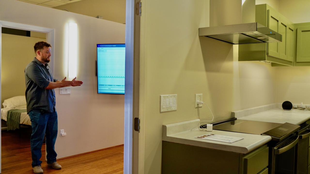 Through a kitchen doorway, Keith Graeber stands talking in living room with unseen person. A big screen tv glows blue in background