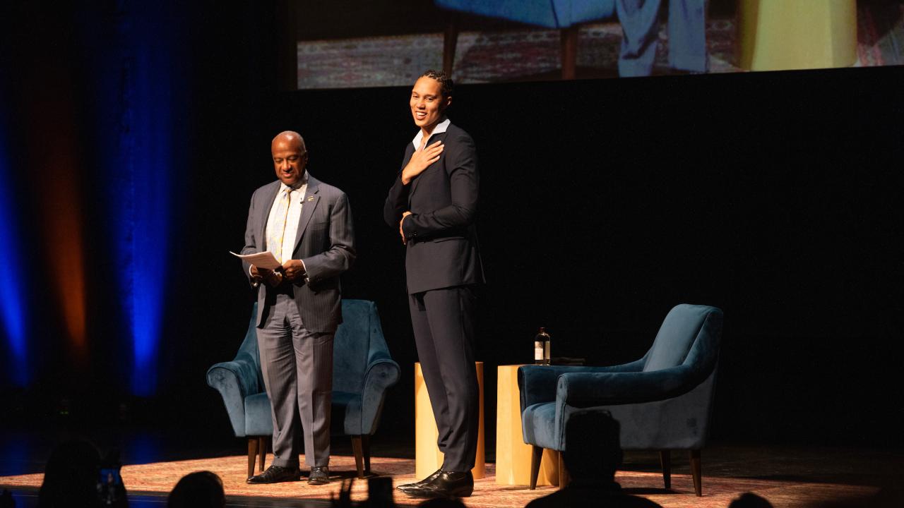 Standing on top of an illuminated theater stage are two people. To the left, a man in a blue suit smiles and look at his notes while on the right a woman in a dark blue suit is standing and pressing her right hand to her left, honoring the standing ovation she is receiving. The man on the left is Chancellor Gary S. May. The woman on the right is his guest, Brittney Griner. Silhouettes of a standing ovation are present in the bottom of the photo. 