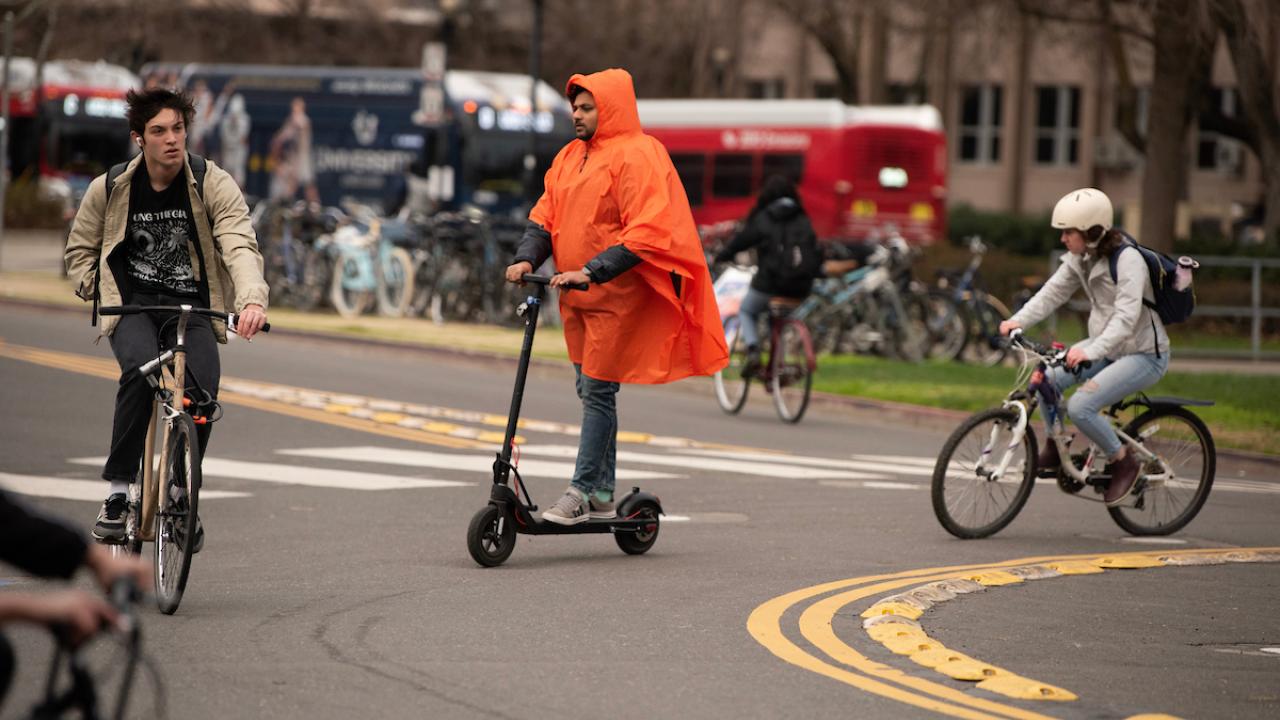 a cyclist in black tee and tan jacket, a man in orange parka and scooter and cyclist in helmet ride around a traffic circle with red Unitrans bus and cars in background