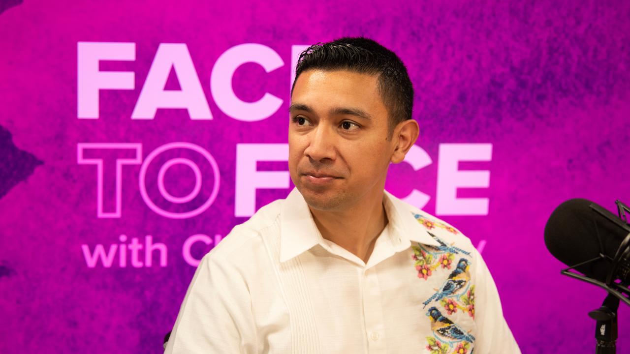 In front of a purple backdrop bearing the Face to Face wordmark logo sits a man in a buttoned white shirt with a design with birds vertically running along its right side. 