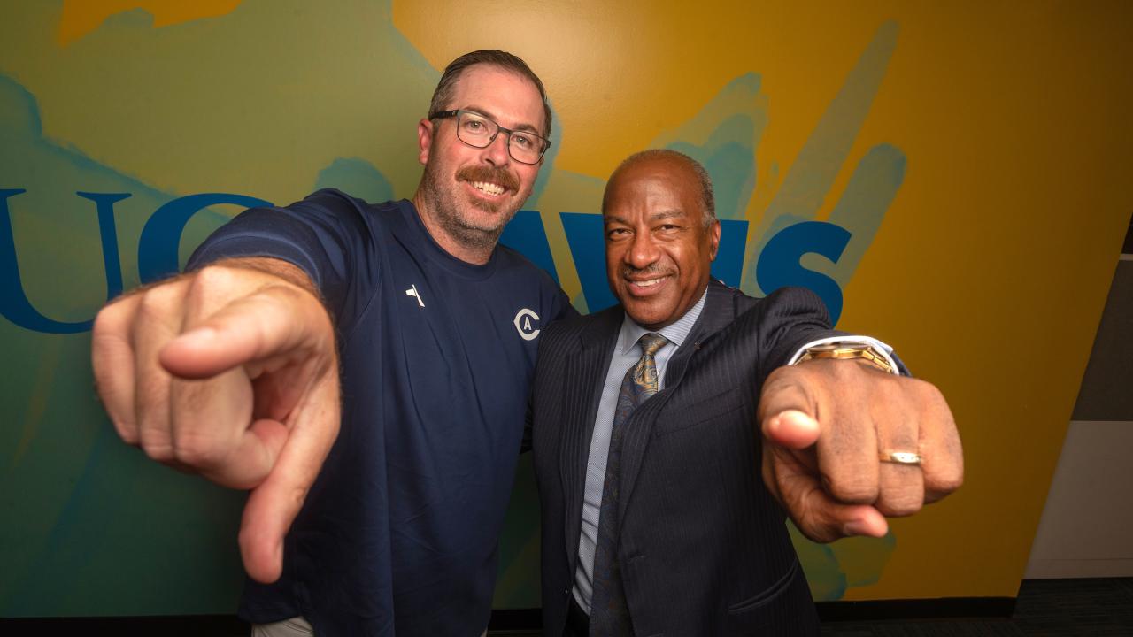 Aggie Football Coach Tim Plough and Chancellor Gary S. May point at the camera in energetic fashion while taking a photo in front of a wall with a UC Davis logo and gradient pattern. 
